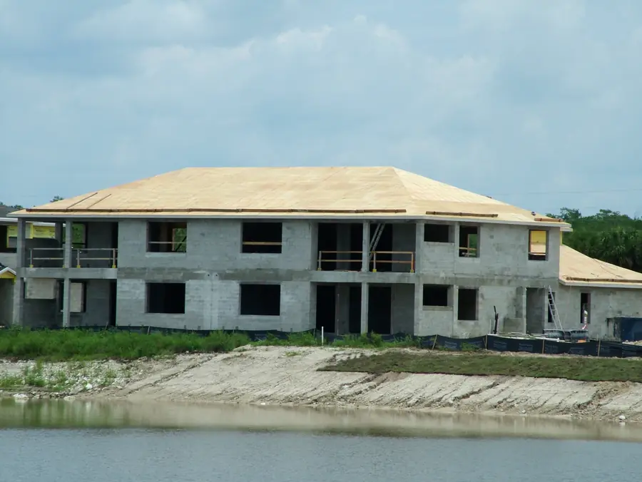 A two-storey concrete block building under construction beside a lake, roof sheathing on and windows not yet in