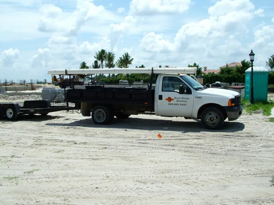 A white South Florida Plumbing F-350 flatbed with PVC pipe on its rack, parked on a sandy construction site; the door reads South Florida Plumbing, CFC041717 and the office number, (239) 643-2431